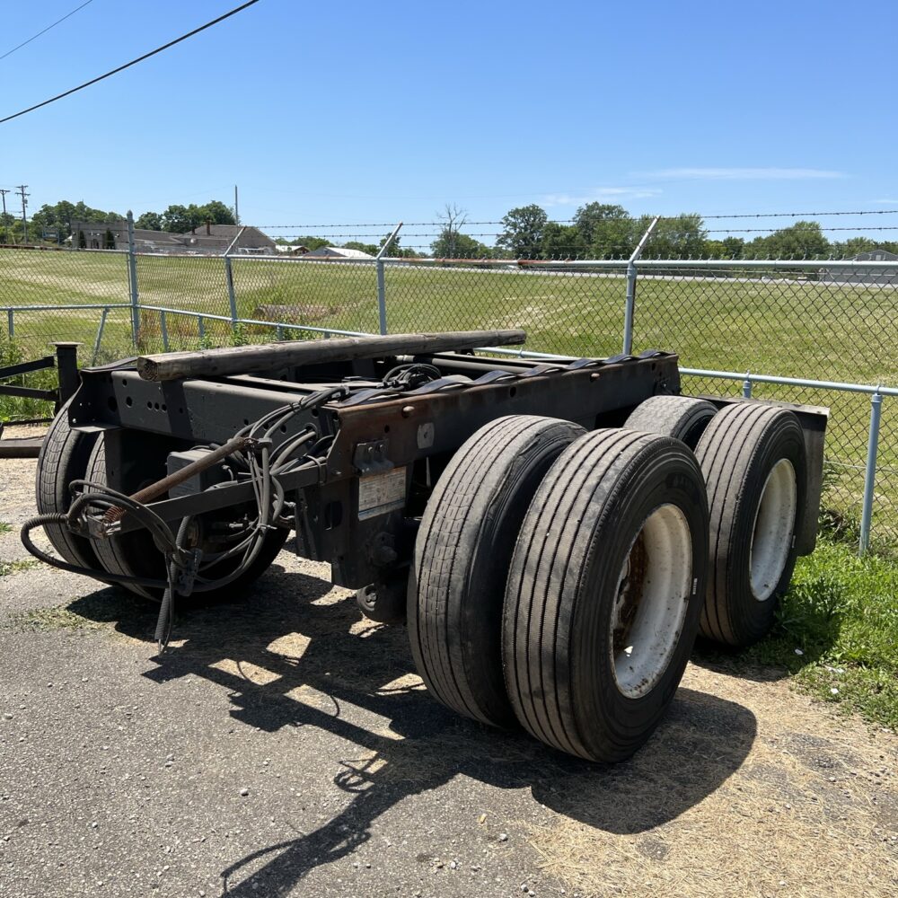 Rear Tandem Axel Assembly, showing 6 tires parked outdoors on a paved surface near a chain-link fence under a clear blue sky