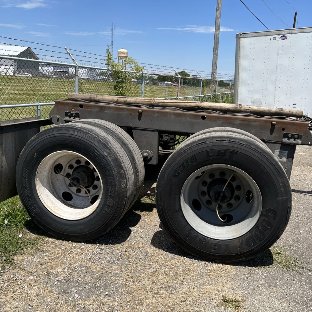 Close-up side view of a semi-truck's rear tandem axle assembly, featuring two large Goodyear tires, parked on a gravel and dirt surface next to a chain-link fence under a clear sky.