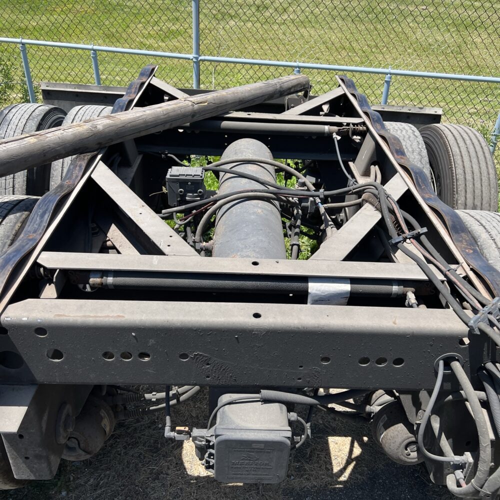 Top-down view of a semi-truck's rear tandem axle assembly, showing the driveshaft, suspension, and other mechanical components, with a chain-link fence and a red SUV visible in the background.