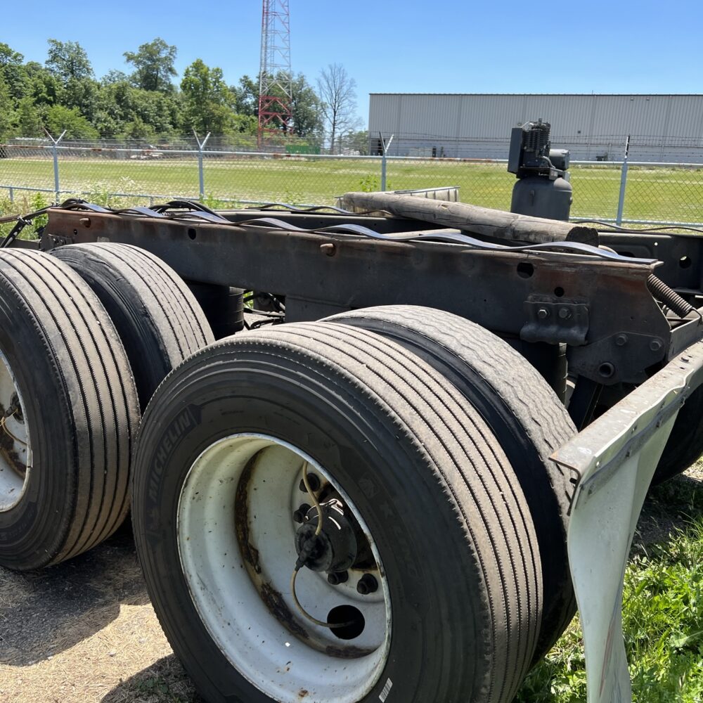 Close-up of a truck's rear axle with dual Michelin tires parked outdoors near a fenced industrial area, showcasing heavy-duty wheels and undercarriage components.