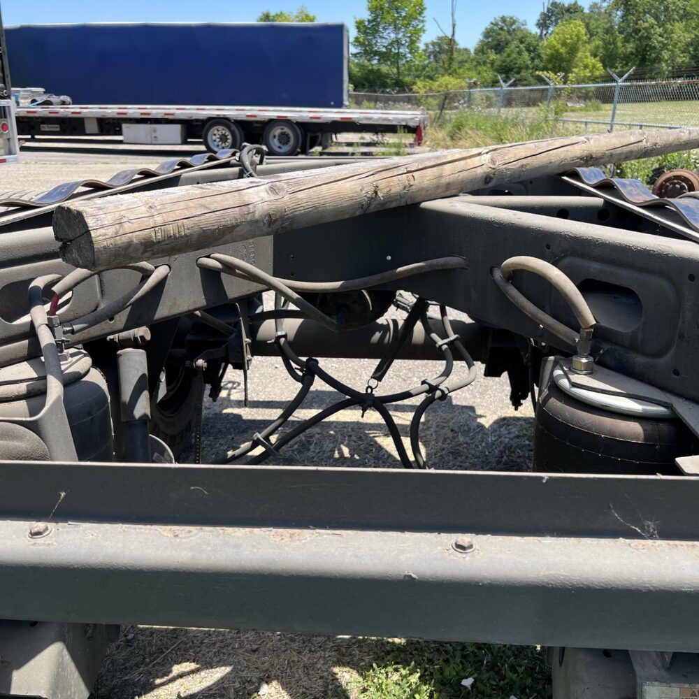 Rear view of a truck chassis showing suspension components, air bags, and a wooden beam resting on top, with a flatbed trailer in the background at an outdoor industrial site.