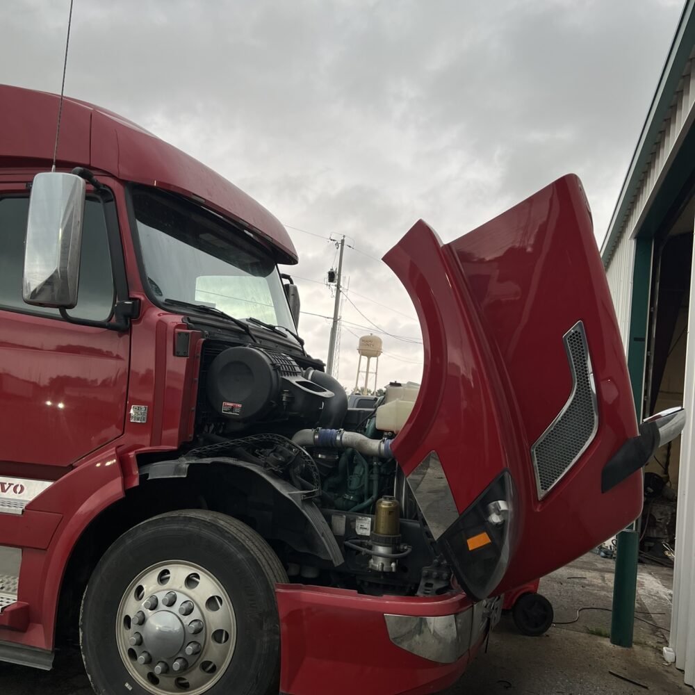 red Volvo semi truck with its hood raised, showing the engine area, parked in front of a repair shop on a cloudy day.