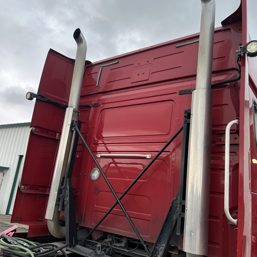 Rear of red Volvo semi truck with dual exhaust stacks and frame supports near repair equipment.