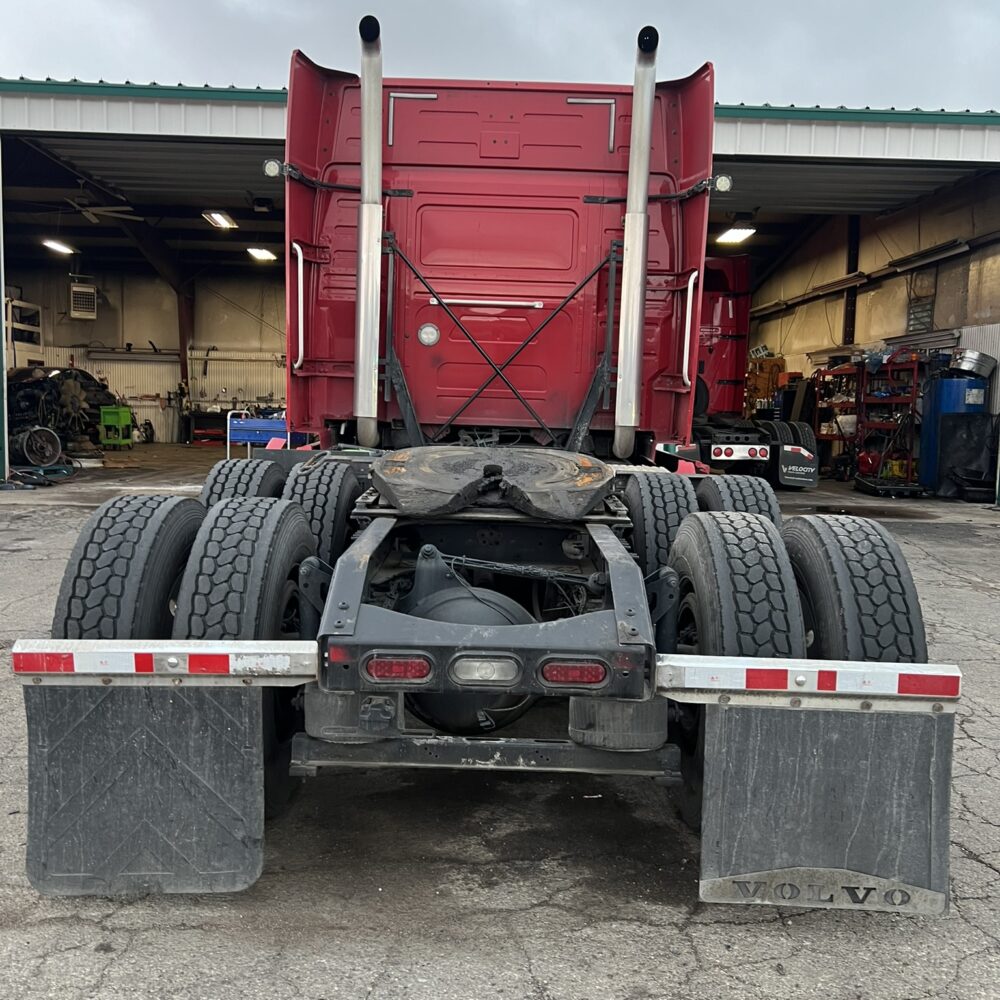 Rear of red Volvo semi truck with dual exhaust stacks, mud flaps, and tires, parked outside a repair shop.