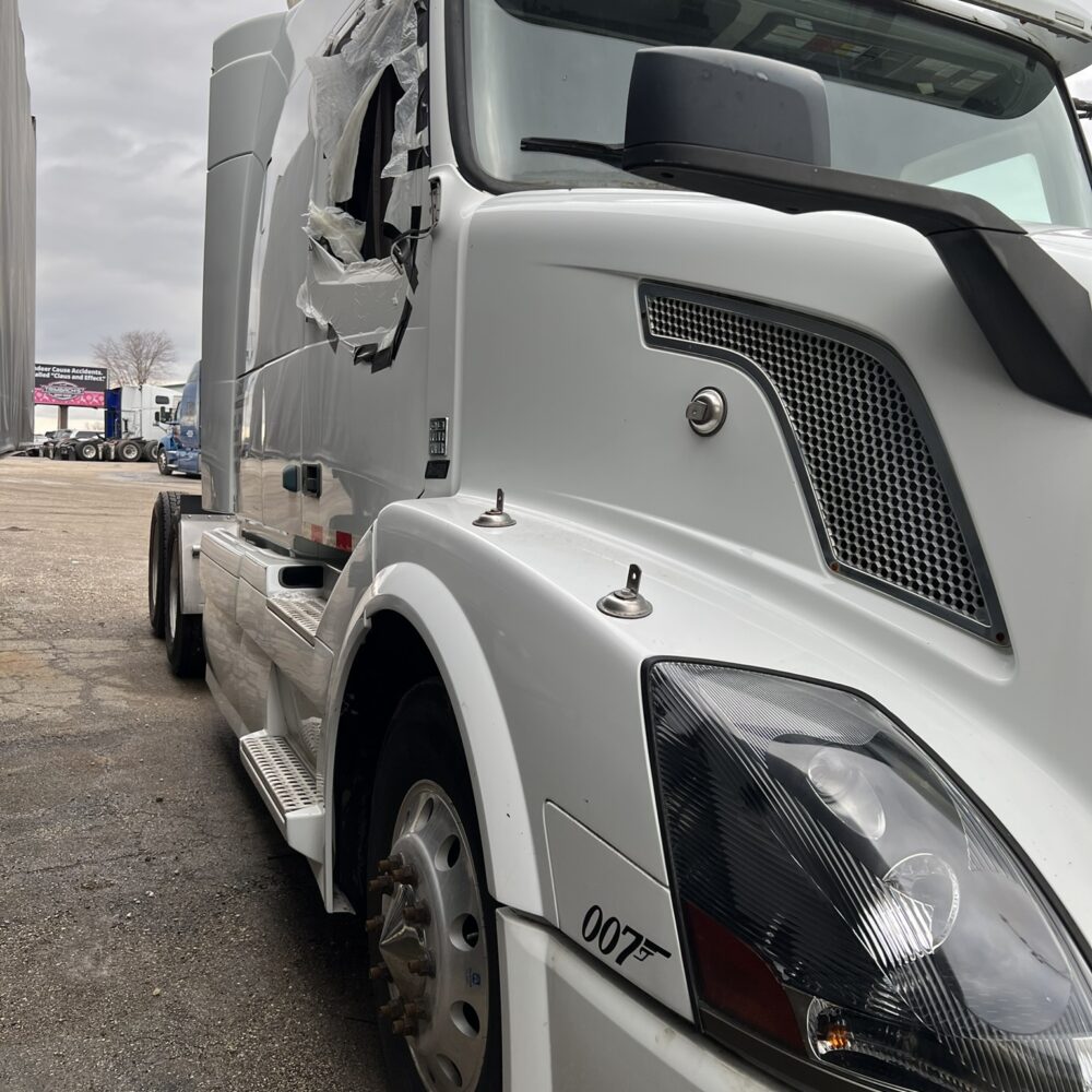 Damaged side of a white Volvo semi truck with taped-up window.